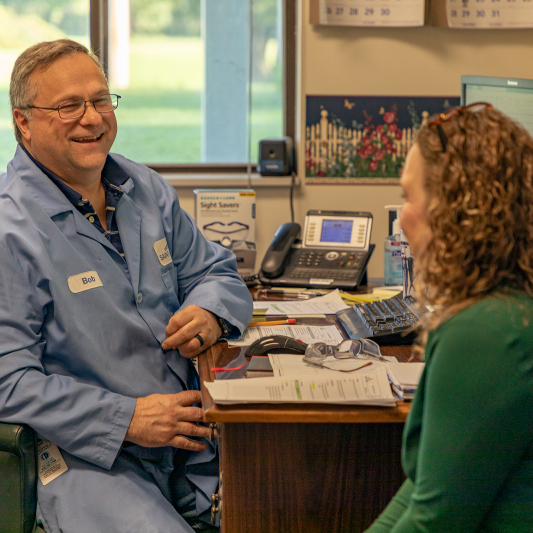 A man sits at his desk while speaking with a female coworker