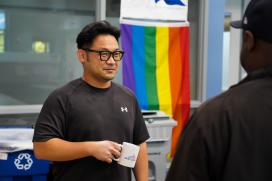 An Asian man stands in an office speaking with a colleague while holding a coffee mug. Behind him hangs a rainbow flag.