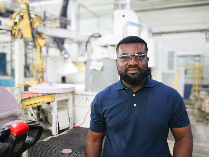 A Black man wearing a blue polo shirt stands in a factory setting in front of white machinery
