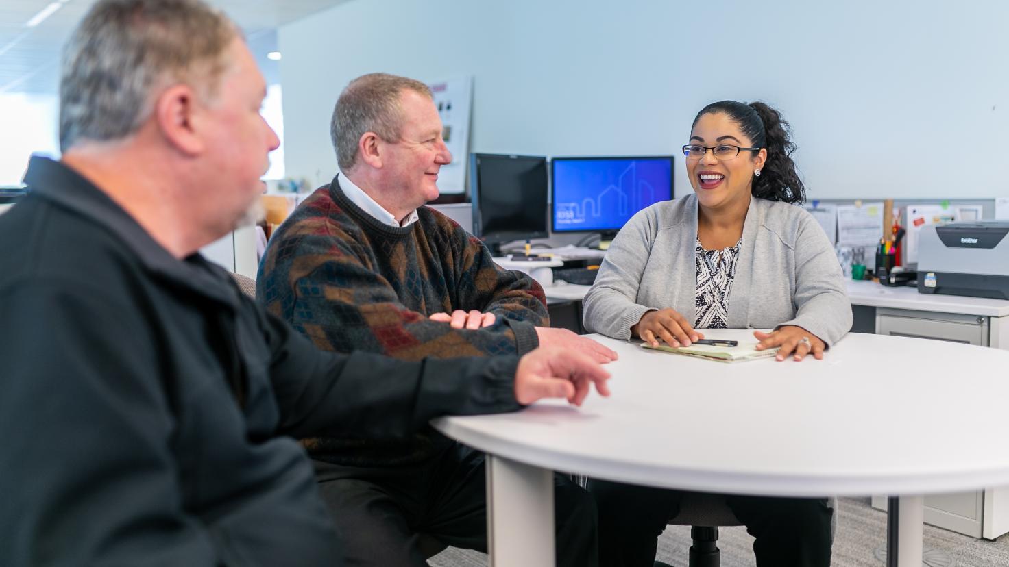 Two men and one woman seated around a round table in an office setting 